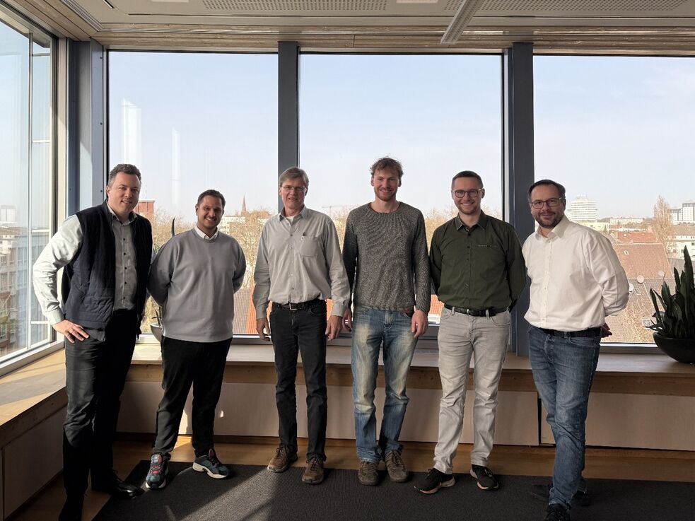 Sechs Männer stehen in einer Reihe in einem Büro mit großen Fenstern und Blick auf die Stadt.